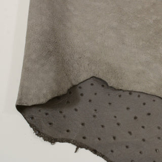 Detail photo of a piece of earl grey colored lambskin with a manufactured ostrich-like pockmark texture. Image shows folded over section that represents both sides, including a lighter-grey, smooth backside.